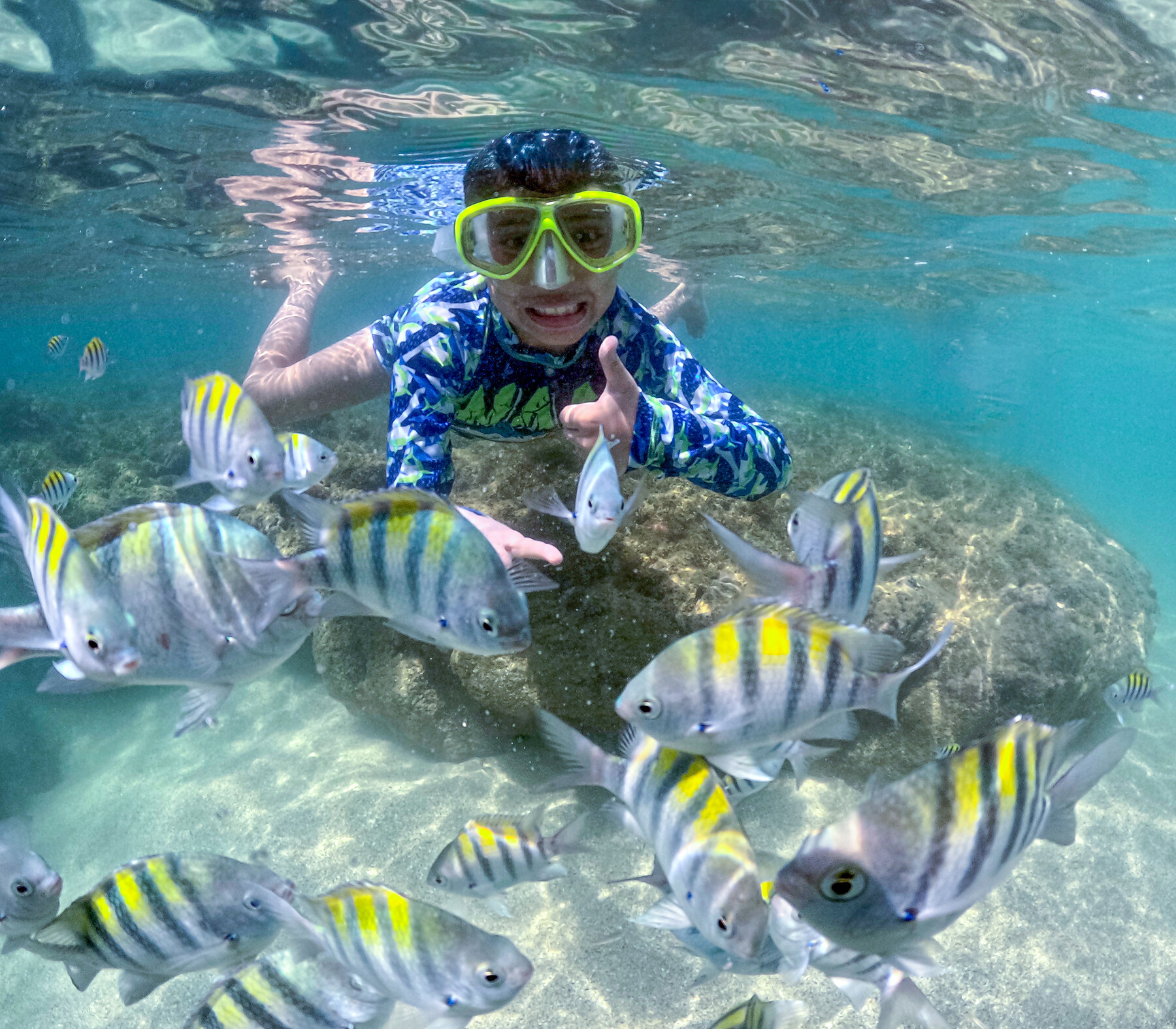 Mergulhador com máscara sorrindo cercado de peixes listrados nas piscinas naturais de Maceió
