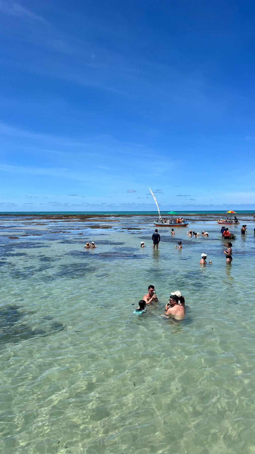 Turistas nas piscinas rasas de Pajuçara com jangadas ao fundo e céu azul