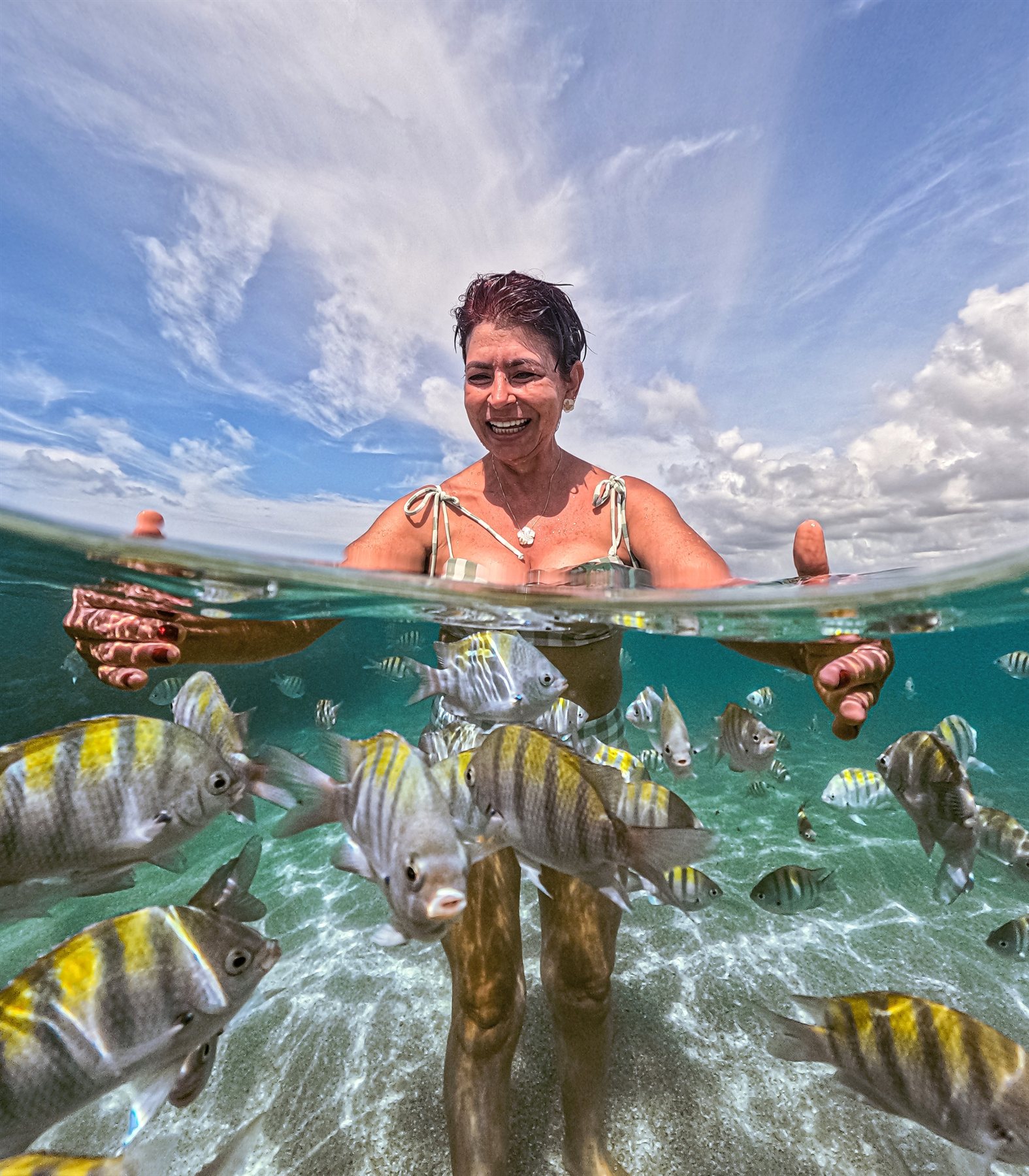 Banhista sorrindo cercada por um cardume de peixes na água cristalina das piscinas naturais de Maceió
