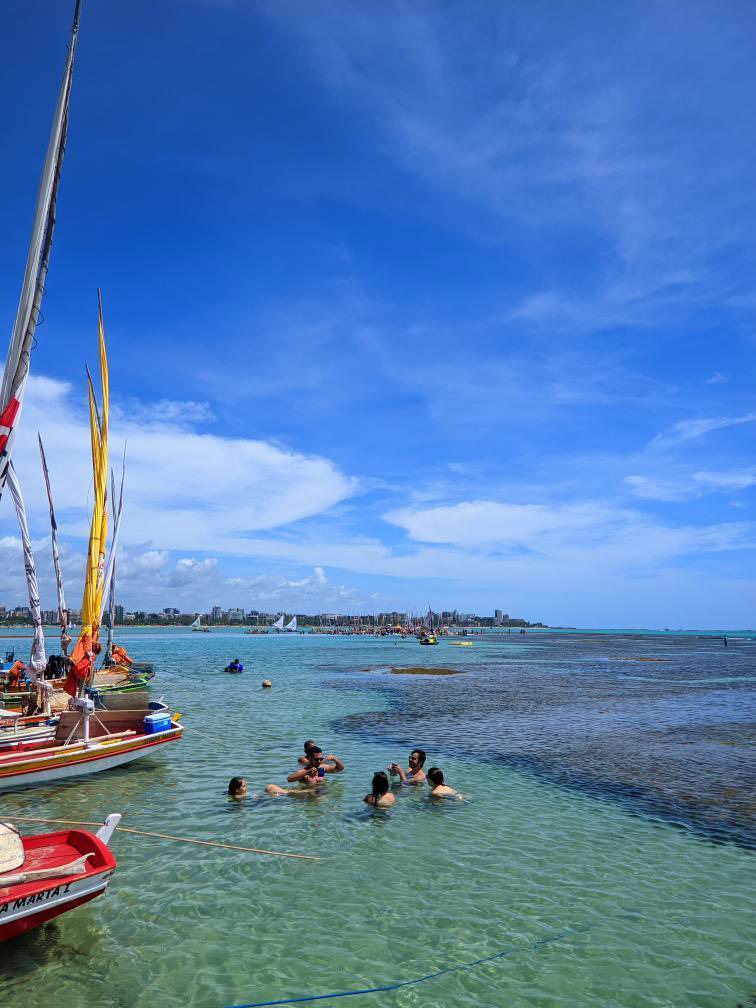 Turistas nas piscinas naturais com jangadas coloridas e a orla de Maceió ao fundo