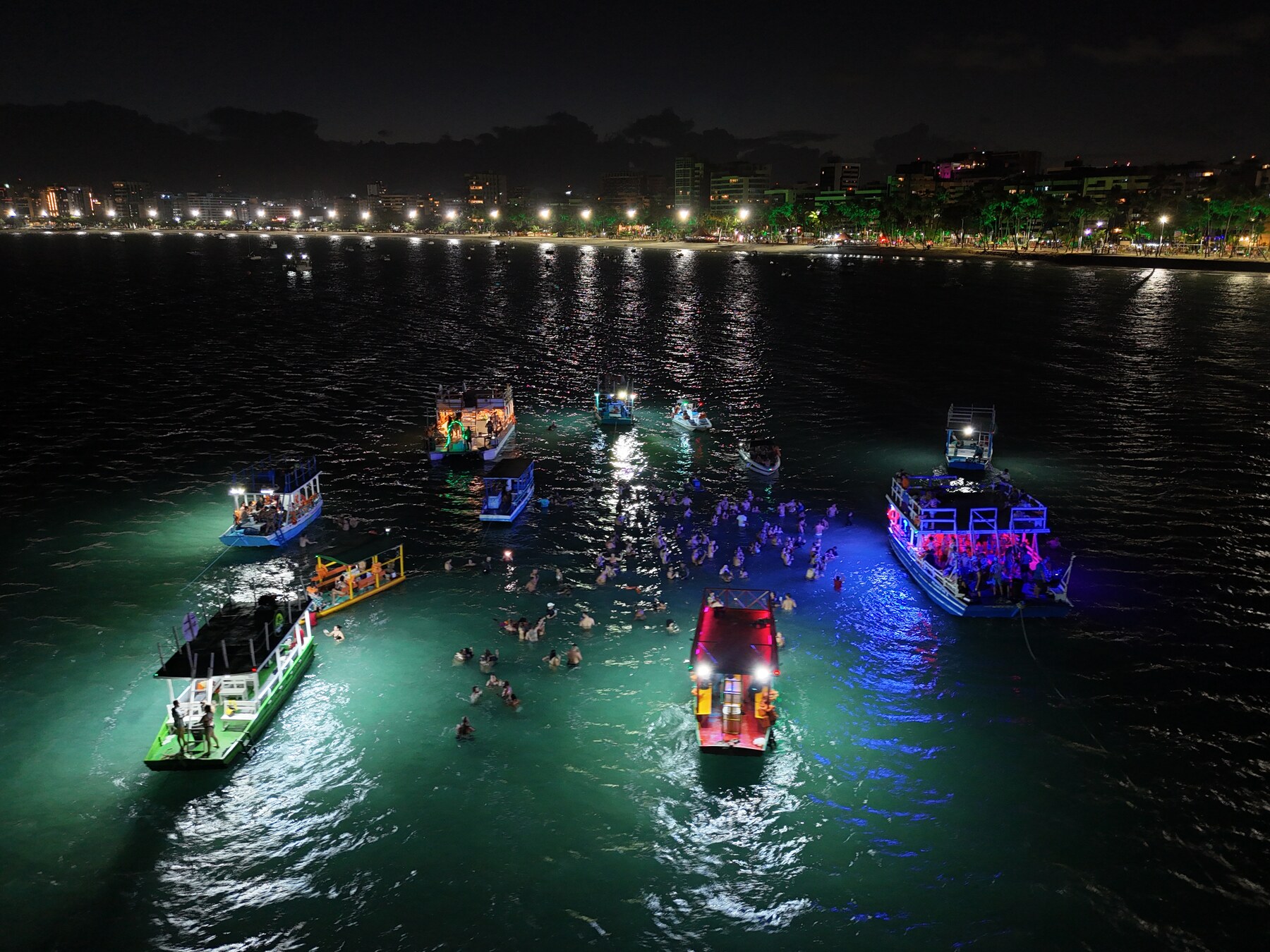 Vista aérea do banho de lua na Ponta Verde — barcos iluminados e dezenas de pessoas na água à noite