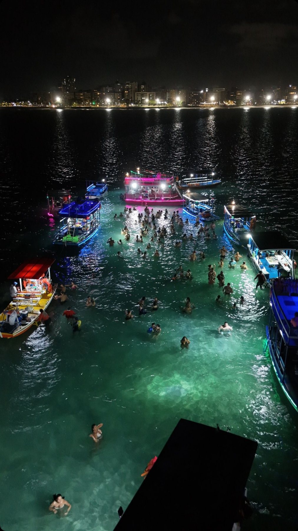 Banho de lua à noite com água turquesa iluminada, barcos coloridos e a orla de Maceió ao fundo